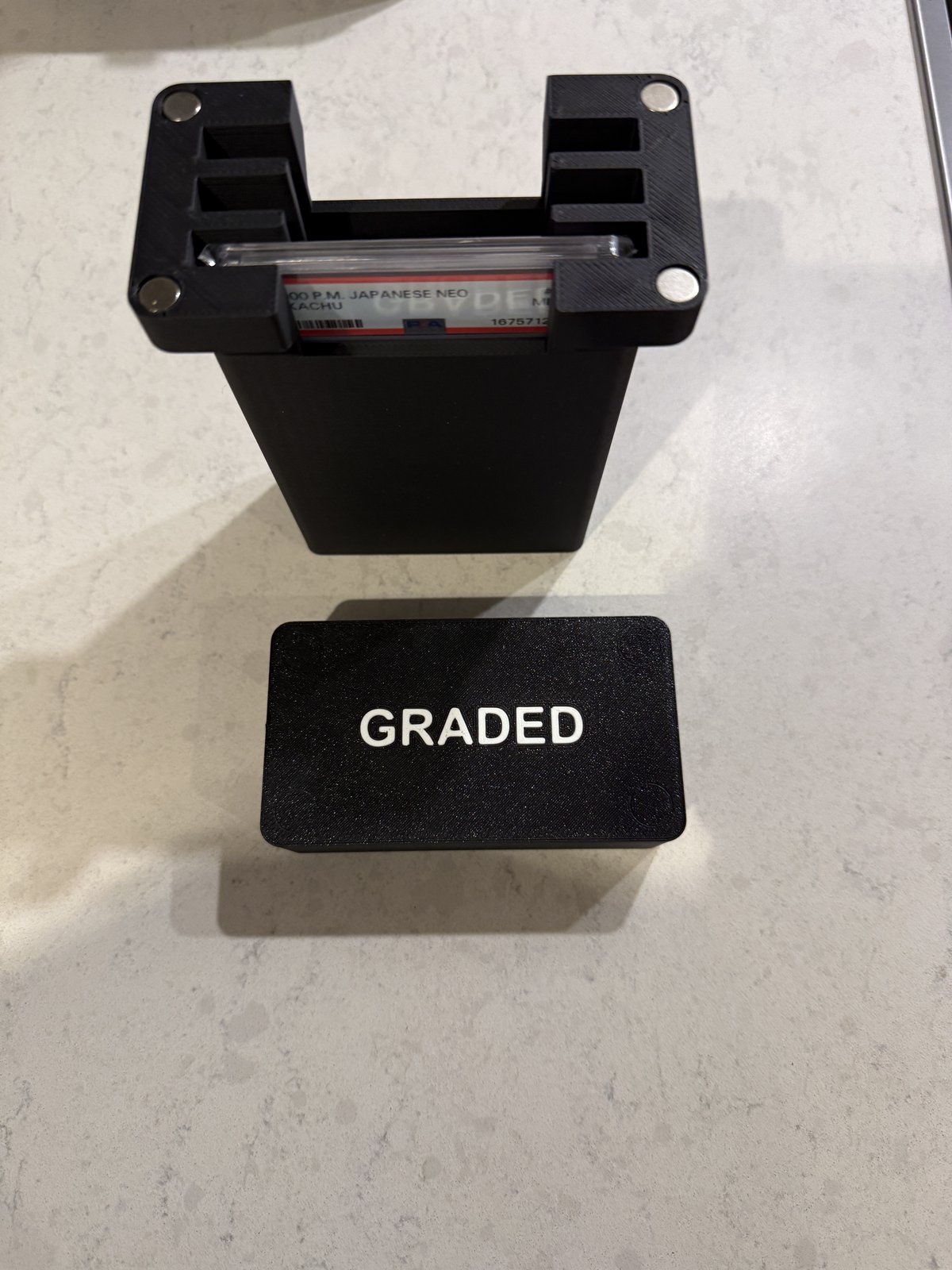 A real Formada3D-printed graded card case, photographed on a kitchen counter, with a PSA-graded Pikachu card seated in the open case next to the matching closed GRADED lid case