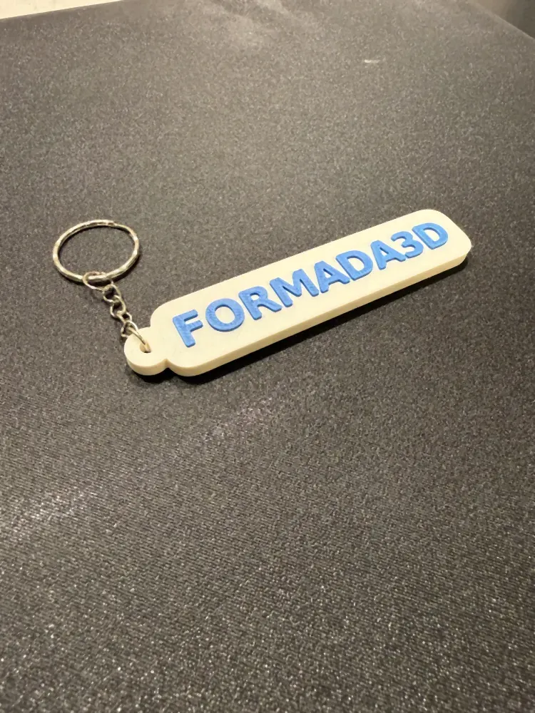 A white FORMADA3D keyring tag with raised blue lettering and a split ring, on a dark bench