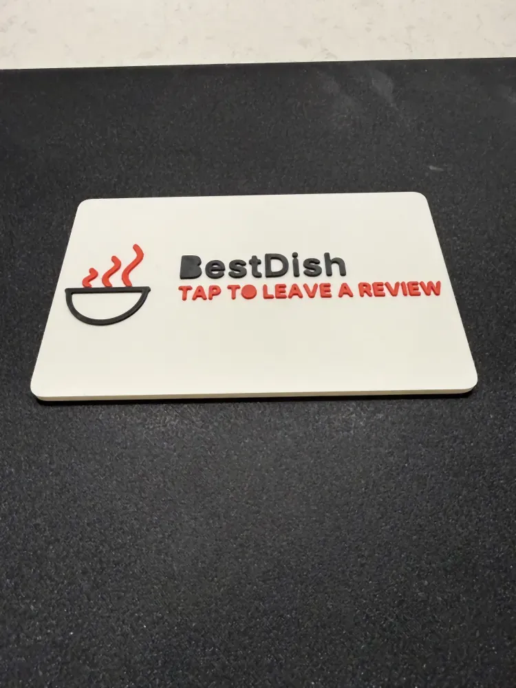 A white printed counter plaque with a restaurant logo and the words TAP TO LEAVE A REVIEW in raised colour